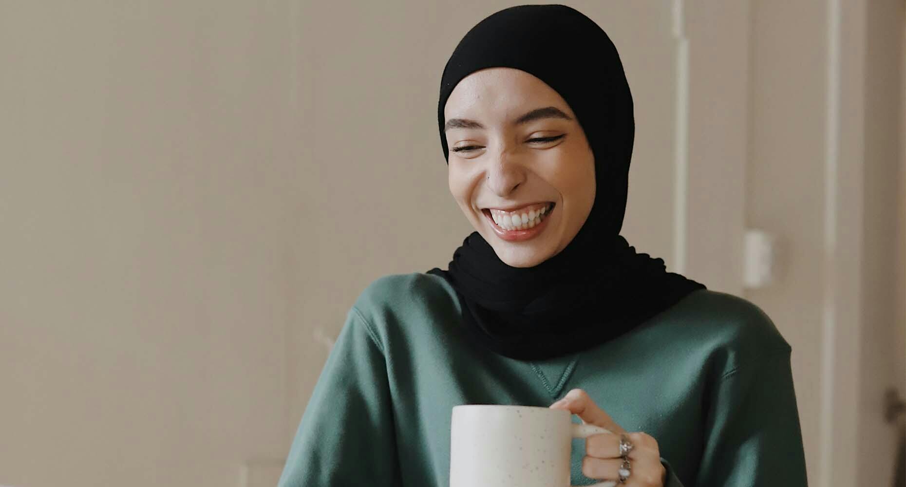 A woman wearing a headscarf enjoying a cup of coffee and laughing.