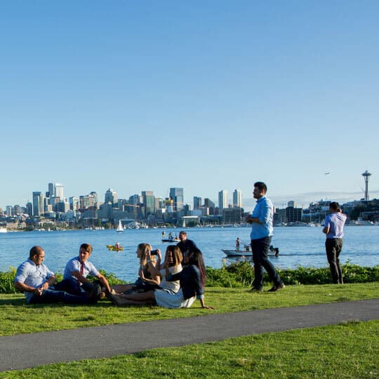 company employees in work clothes taking a break, sitting on the grass by water with buildings in the background